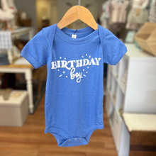 Load image into Gallery viewer, A blue short-sleeve baby bodysuit hanging on a wooden hanger, featuring the words 'BIRTHDAY boy' in white script and block lettering with small star accents. The bodysuit is photographed in a softly blurred boutique setting with wooden floors and display shelves in the background.