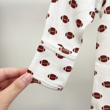 Load image into Gallery viewer, A close-up of a baby garment sleeve featuring optional foldover cuffs with a brown football pattern, on white pointelle fabric, held by a hand against a neutral light gray back