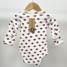 Load image into Gallery viewer, The back view of a white long-sleeve baby bodysuit with an all-over brown football print, hanging on a wooden hanger against a light gray background. A paper brand tag reading 'Grace Lou' is attached to the neckline.