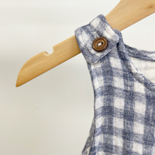 Load image into Gallery viewer, Close up details of the strap on a blue gingham, baby boy jumpsuit on a wooden hanger in front of a white background.