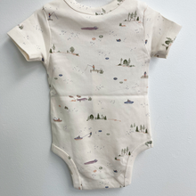 Load image into Gallery viewer, A back view of a short-sleeve baby bodysuit in a warm cream color, featuring a delicate all-over print of small boats, trees, and lake scenes in muted green, blue, and brown tones.
