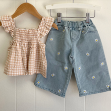 Load image into Gallery viewer, Light wash blue denim pants with white daisy flowers on a hanger with a peach gingham shirt styled beside it.