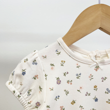 Load image into Gallery viewer, A close up picture of the necklinen of a floral, organic cotton, baby dress that also shows a cap sleeve with elastic and the round neckline adorned with a decorative ribbon bow.