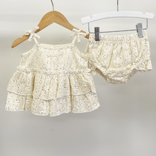 Load image into Gallery viewer, A two-piece baby girl's outfit set consisting of a tiered ruffle tank top and matching bloomers, both in a cream-colored cotton fabric with a tiny white floral print. The tank top is displayed on a wooden hanger, and the matching bloomers are held by silver metal clips on a white hanger, set against a plain light gray background.