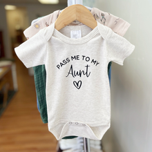 Load image into Gallery viewer, A light heather gray baby bodysuit hanging on a wooden hanger, featuring the black printed text 'PASS ME TO MY Aunt' and a heart. Other baby clothes are partially visible behind it on a store display.