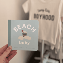 Load image into Gallery viewer, Book titled 'Beach Baby' held in front of a Long Live Boyhood t shirt with a blurred background