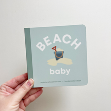 Load image into Gallery viewer, Hand holding a children's book titled 'Beach Baby' with a sandcastle illustration on a light gray background.