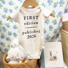 Load image into Gallery viewer, Baby onesie with 'First edition published 2026' text on a hanger against a floral background