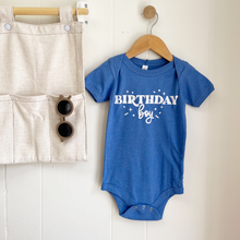 Load image into Gallery viewer, A royal blue baby bodysuit with 'BIRTHDAY boy' printed in white on the front, hanging on a wooden hanger against a white paneled wall. A beige linen pocket organizer with sunglasses is hanging to the left as a styled prop.