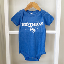 Load image into Gallery viewer, A royal blue short-sleeve baby bodysuit hanging on a wooden hanger, featuring the words 'BIRTHDAY boy' in white across the chest, set against a white wooden frame background.