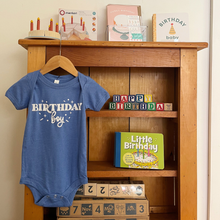 Load image into Gallery viewer, A blue baby bodysuit with 'BIRTHDAY boy' printed in white on the front, hanging on a wooden hanger in front of a wooden bookshelf filled with baby-themed birthday items.