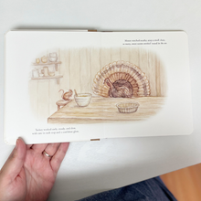 Load image into Gallery viewer, A hand holding an open children's picture book showing a watercolor illustration of a turkey cooking in a kitchen, with printed text on both pages, perfect for Thanksgiving traditions with children.