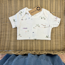 Load image into Gallery viewer, A person admiring a short-sleeve baby bodysuit in a warm cream color, featuring a delicate all-over print of small boats, trees, and lake scenes in muted green, blue, and brown tones folded in half in a wicker basket.