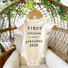 Load image into Gallery viewer, Baby onesie with 'First edition published 2026' text on a hanger with greenery