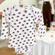 Load image into Gallery viewer, A white long-sleeve baby bodysuit with an all-over brown football print, hanging on a white hanger in a softly blurred inside the Gracie Lou children's boutique setting.