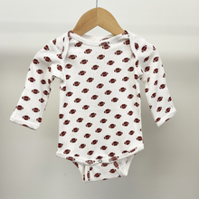 Load image into Gallery viewer, A baby long-sleeve bodysuit in white with an all-over brown football print, on pointelle cotton fabric, hanging on a wooden hanger against a plain light gray background.