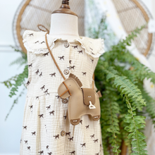 Load image into Gallery viewer, Details of the bodice of a toddler size muslin dress with a horse print on a cream dress styled on a mannequin in front of a green fern inside a children's boutique, Gracie Lou.