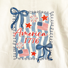 Load image into Gallery viewer, A close up of a toddler girls tee shirt with a patriotic design including American flags, stars, bows, and fireworks on a white background with 'America 1776' text.