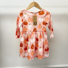 Load image into Gallery viewer, A pink long-sleeve baby dress with an orange and white pumpkin print, hanging on a wooden hanger against a plain light gray wall. The dress has a gathered waist and a paper tag from the brand 'Gracie Lou'.
