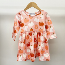 Load image into Gallery viewer, A toddler girl's long-sleeve dress with a pumpkin print, hanging on a wooden hanger against a plain light gray background. The dress has a light pink base with orange, red, and white pumpkins printed all over it.