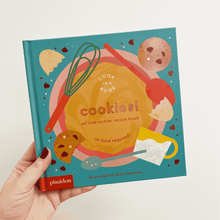 Load image into Gallery viewer, A hand holding an interactive cook book for kids with a cookie recipe inside.