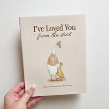 Load image into Gallery viewer, A hand holding a hardcover children's book titled "I've Loved You from the start" against a plain light background. The cover shows a beige-toned illustration of a duck and duckling with brown and white coloring.