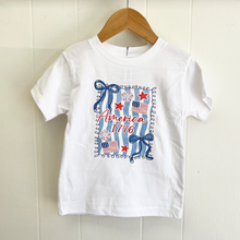 Load image into Gallery viewer, White toddler girls t-shirt with patriotic design, including bows and flags, on a wooden hanger against a white wall.