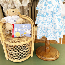 Load image into Gallery viewer, Teddy bear in a wicker chair with a book titled 'Little America' next to a floral dress on a mannequin.