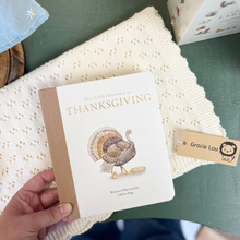 Load image into Gallery viewer, A hand holds a children's book titled "Our Little Adventure at Thanksgiving" with a cream cover and kraft paper spine, resting on a cream textured blanket over a green surface. A small paper tag reading "Gracie Lou" is visible beside it.