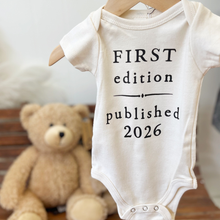 Load image into Gallery viewer, Baby onesie with 'First edition published 2026' text, hanging on a hanger with a teddy bear in the background.