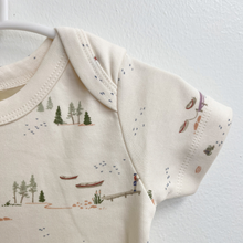 Load image into Gallery viewer, Close up details of the sleeve of a short-sleeve baby bodysuit in a warm cream color, featuring a delicate all-over print of small boats, trees, and lake scenes in muted green, blue, and brown tones.