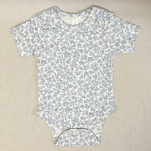 Load image into Gallery viewer, Baby onesie with blue floral pattern on a beige fabric background