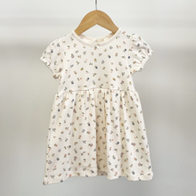 Load image into Gallery viewer, A floral baby dress on a wooden hanger, the dress is a creamy white with multi color floral stems in an all-over pattern.