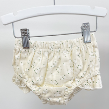 Load image into Gallery viewer, A pair of baby bloomers in a cream-colored cotton fabric with a tiny white floral print, hanging from a white plastic hanger with metal clips against a plain light gray background.