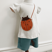 Load image into Gallery viewer, Shell colored A-line t-shirt with a ladybug crossbody purse and Laguna (teal) shorts on a plain background