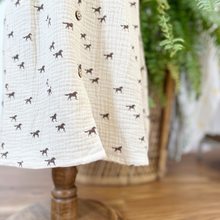 Load image into Gallery viewer, A close up of the skirt detail on a toddler size muslin dress with a horse print on a cream dress styled on a mannequin in front of a green fern.